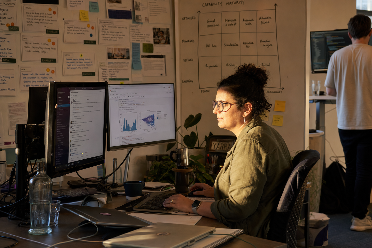 A data engineer in her thirties sits at a dual-monitor workstation in a sun-warmed Australian tech office, screens showing a Jupyter notebook and a chat thread, with a sprint board and whiteboard visible behind her.